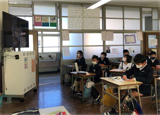 現場中継をモニターで真剣に見ている児童